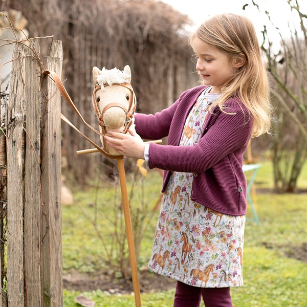 Mädchen im Pferdekleid und Strickjacke von Enfant Terrible spielt mit Steckenpferd auf einer Wiese